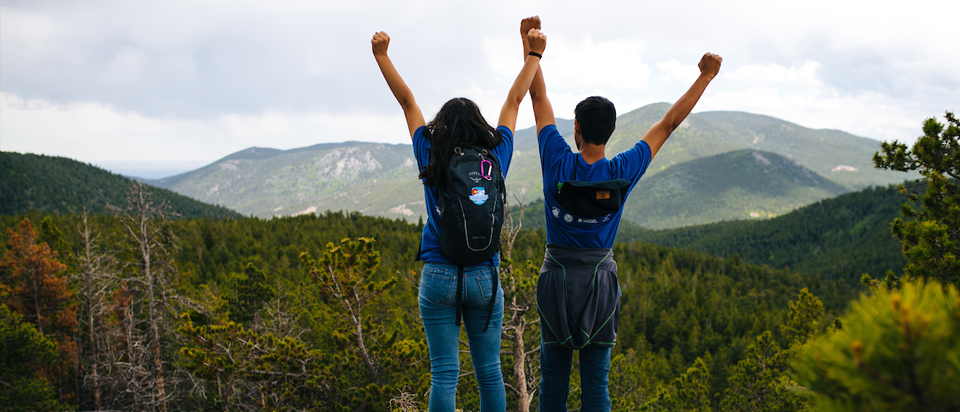 Image of two young people posing with their arms up at the edge of a cliff with a mountain view.