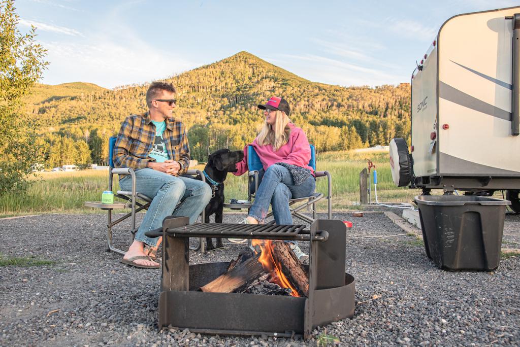 Man and female with black dog sit behind campfire with mountains in the background