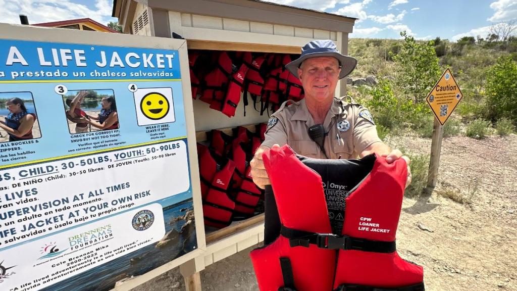 A park ranger standing in front of a free day-use life jacket station holds a red life jacket in front of him