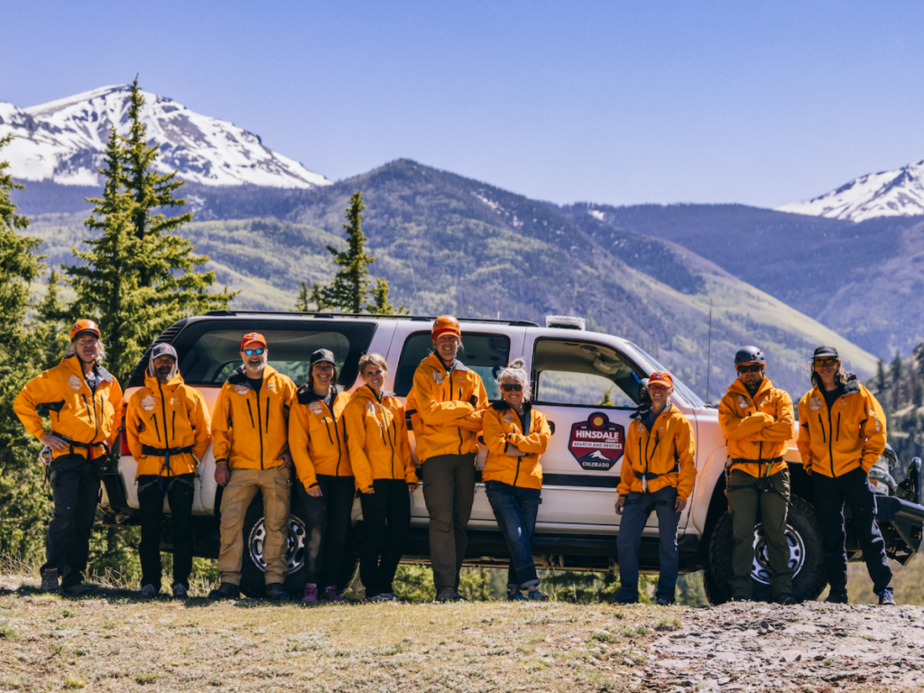 Picture of the Hinsdale County Search and Rescue unit wearing orange jackets and a mountain landscape in the background