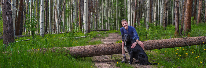 A hiker poses against a forest backdrop with a dog